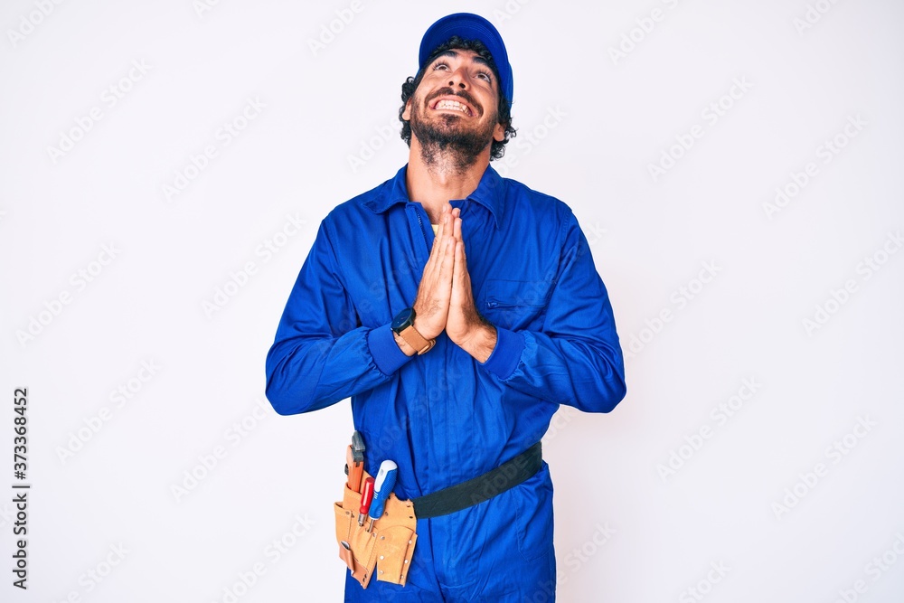 Handsome young man with curly hair and bear weaing handyman uniform begging and praying with hands together with hope expression on face very emotional and worried. begging. Handsome young man with curly hair and bear weaing handyman uniform begging and praying with hands together with hope expression on face very emotional and worried. begging.