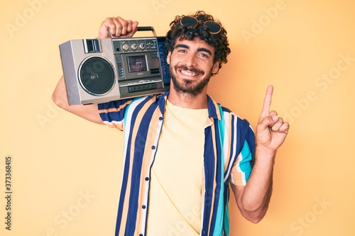 Handsome young man with curly hair and bear holding boombox, listening to music wearing summer look smiling with an idea or question pointing finger with happy face, number one
