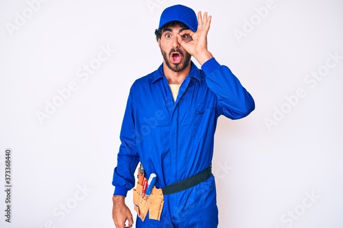 Handsome young man with curly hair and bear weaing handyman uniform doing ok gesture shocked with surprised face, eye looking through fingers. unbelieving expression.