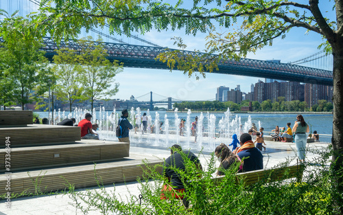 Fototapeta Naklejka Na Ścianę i Meble -  Brooklyn, NY / USA - 8/20/20: Landscape view of Domino Park's fountain and seating steps. The East River and Williamsburg Bridge in the distance.