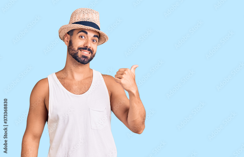 Young hispanic man wearing casual summer hat smiling with happy face looking and pointing to the side with thumb up. Young hispanic man wearing casual summer hat smiling with happy face looking and pointing to the side with thumb up.