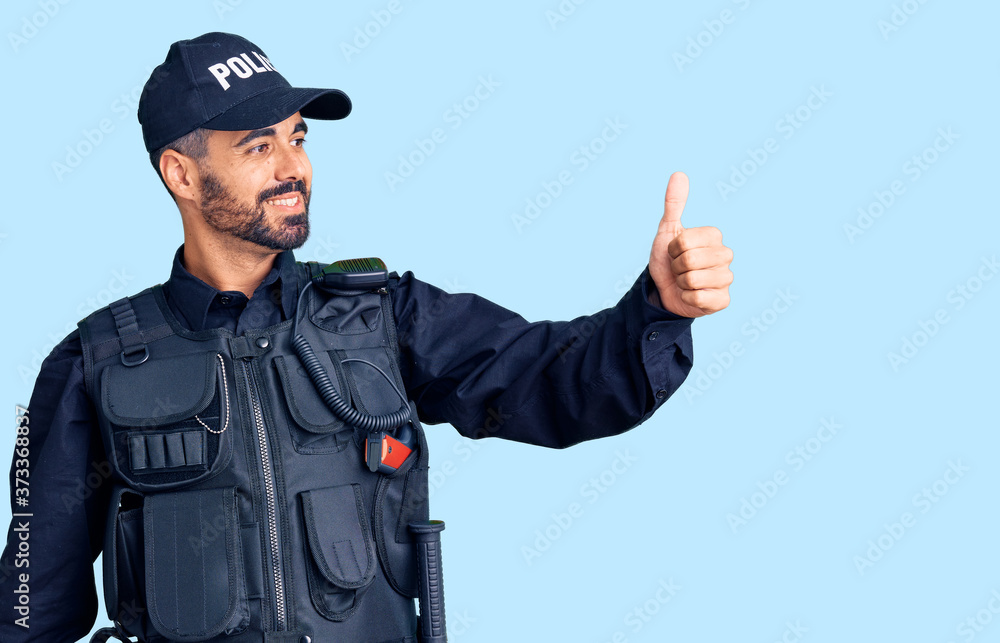 Young hispanic man wearing police uniform looking proud, smiling doing thumbs up gesture to the side Young hispanic man wearing police uniform looking proud, smiling doing thumbs up gesture to the side