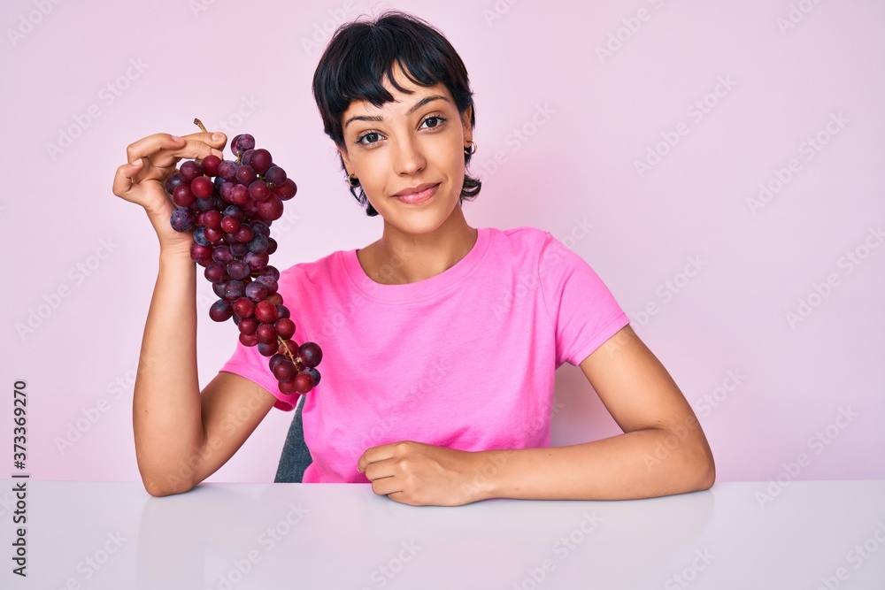 Beautiful brunettte woman holding branch of fresh grapes thinking attitude and sober expression looking self confident Beautiful brunettte woman holding branch of fresh grapes thinking attitude and sober expression looking self confident