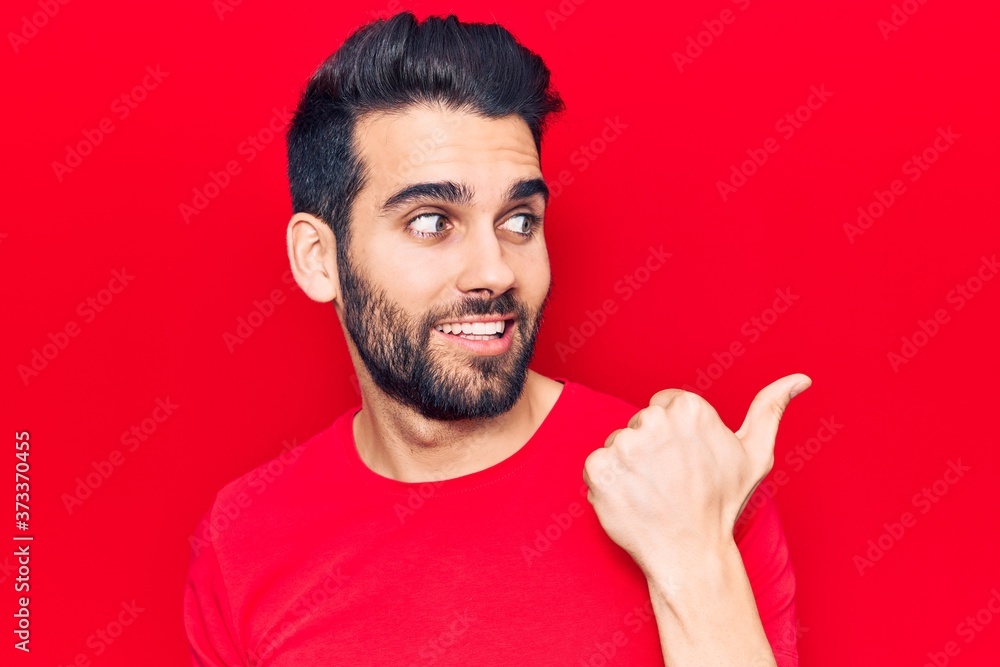 Young handsome man with beard wearing casual t-shirt smiling with happy face looking and pointing to the side with thumb up. Young handsome man with beard wearing casual t-shirt smiling with happy face looking and pointing to the side with thumb up.