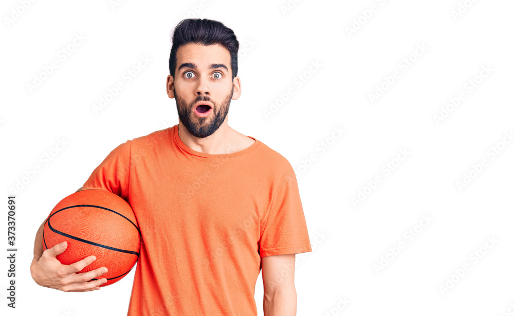 Fototapeta premium Young handsome man with beard hoilding basketball ball scared and amazed with open mouth for surprise, disbelief face