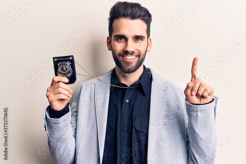 Young handsome man with beard holding police badge surprised with an idea or question pointing finger with happy face, number one © Krakenimages.com