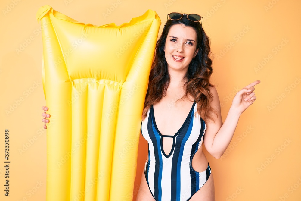 Obraz premium Young beautiful brunette woman wearing swimsuit and holding summer matress float smiling happy pointing with hand and finger to the side