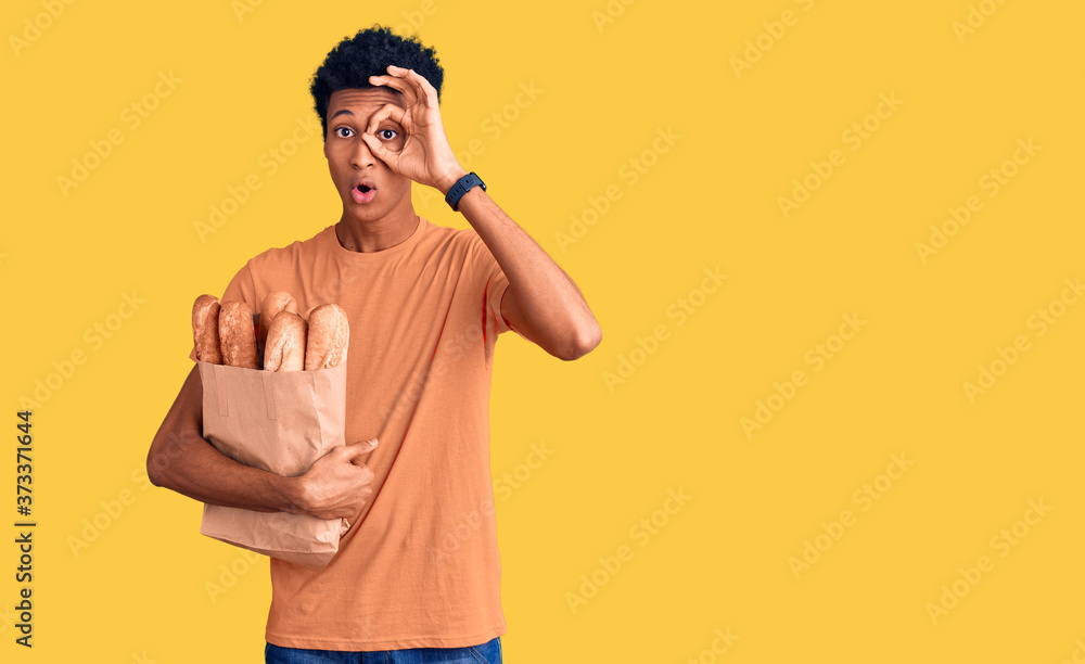 Young african american man holding paper bag with bread doing ok gesture shocked with surprised face, eye looking through fingers. unbelieving expression. Young african american man holding paper bag with bread doing ok gesture shocked with surprised face, eye looking through fingers. unbelieving expression.
