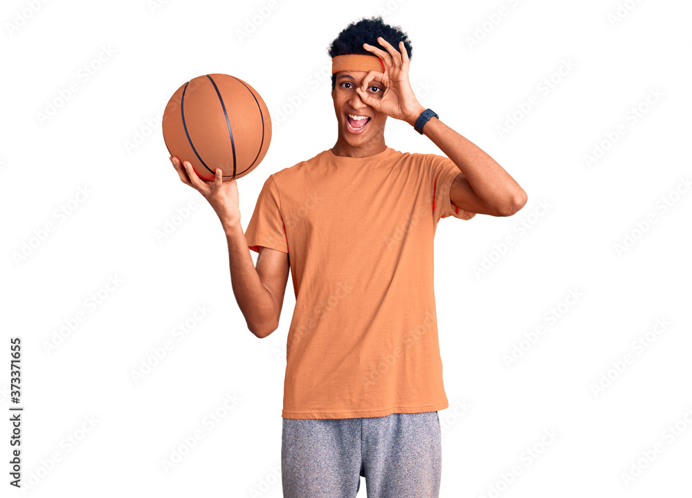Young african american man holding basketball ball smiling happy doing ok sign with hand on eye looking through fingers