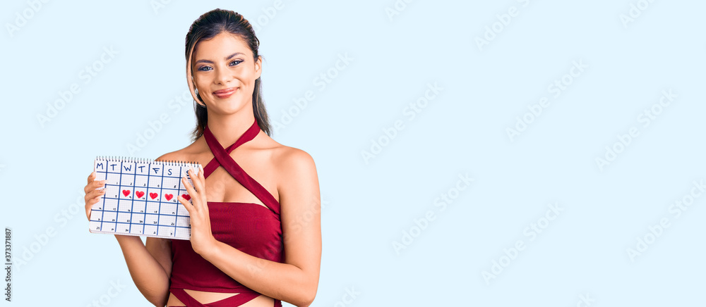 Young beautiful woman holding heart calendar looking positive and happy standing and smiling with a confident smile showing teeth Young beautiful woman holding heart calendar looking positive and happy standing and smiling with a confident smile showing teeth