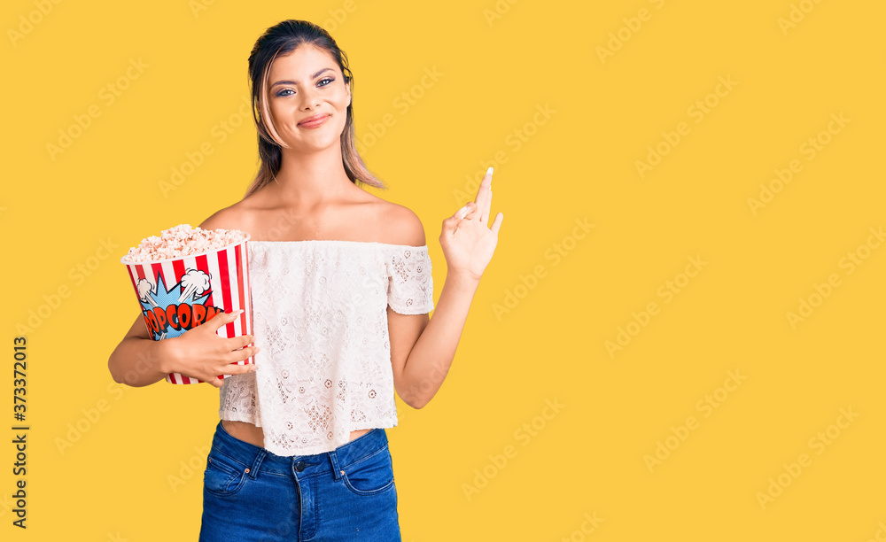 Young beautiful woman holding popcorn doing ok sign with fingers, smiling friendly gesturing excellent symbol Young beautiful woman holding popcorn doing ok sign with fingers, smiling friendly gesturing excellent symbol