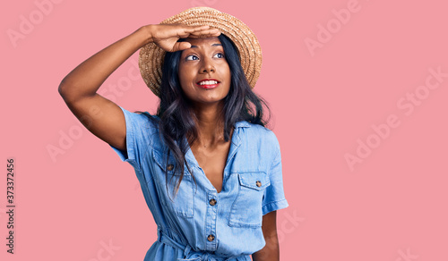 Young indian girl wearing summer hat very happy and smiling looking far away with hand over head. searching concept.