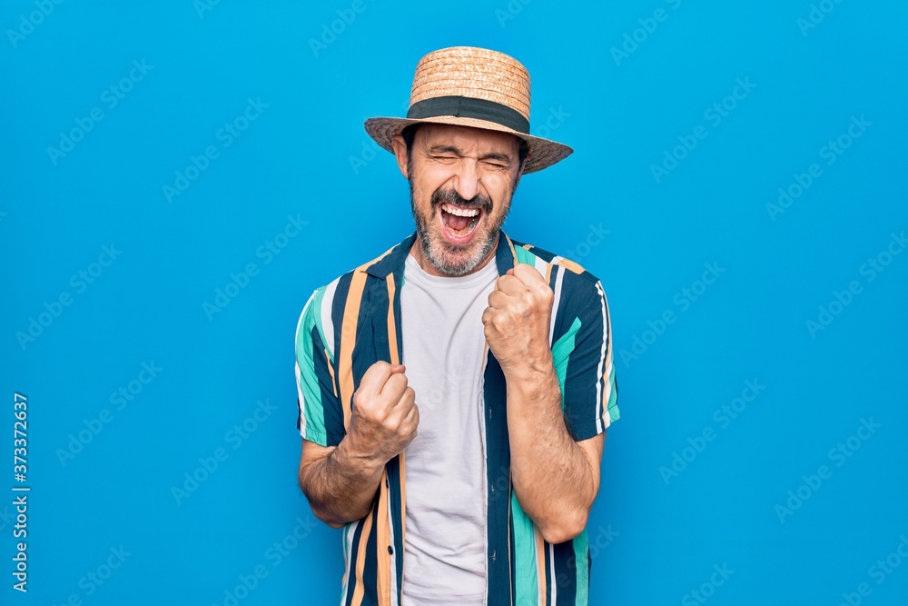 Middle age handsome tourist man on vacation wearing shirt and hat over blue background celebrating surprised and amazed for success with arms raised and eyes closed Middle age handsome tourist man on vacation wearing shirt and hat over blue background celebrating surprised and amazed for success with arms raised and eyes closed