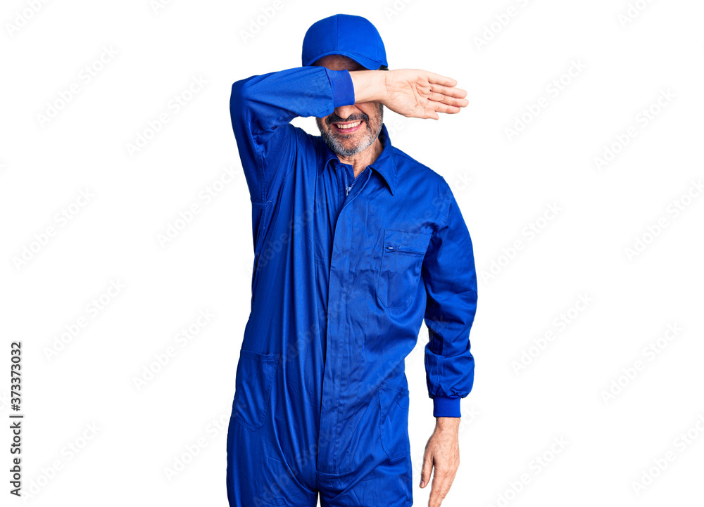Middle age handsome man wearing mechanic uniform covering eyes with arm smiling cheerful and funny. blind concept. Middle age handsome man wearing mechanic uniform covering eyes with arm smiling cheerful and funny. blind concept.
