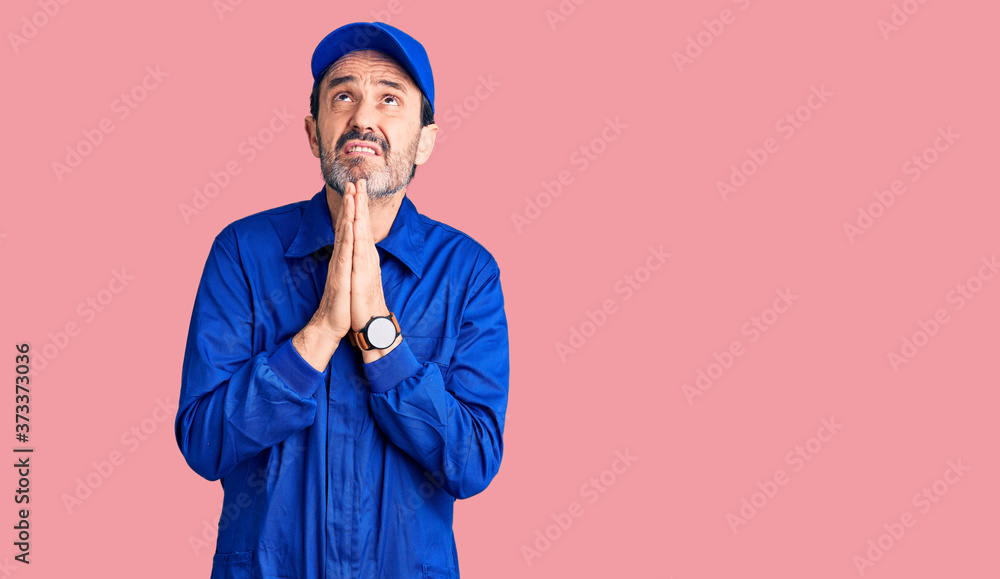 Middle age handsome man wearing mechanic uniform begging and praying with hands together with hope expression on face very emotional and worried. begging. Middle age handsome man wearing mechanic uniform begging and praying with hands together with hope expression on face very emotional and worried. begging.
