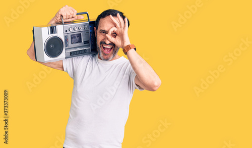 Middle age handsome man listening to music using vintage boombox smiling happy doing ok sign with hand on eye looking through fingers