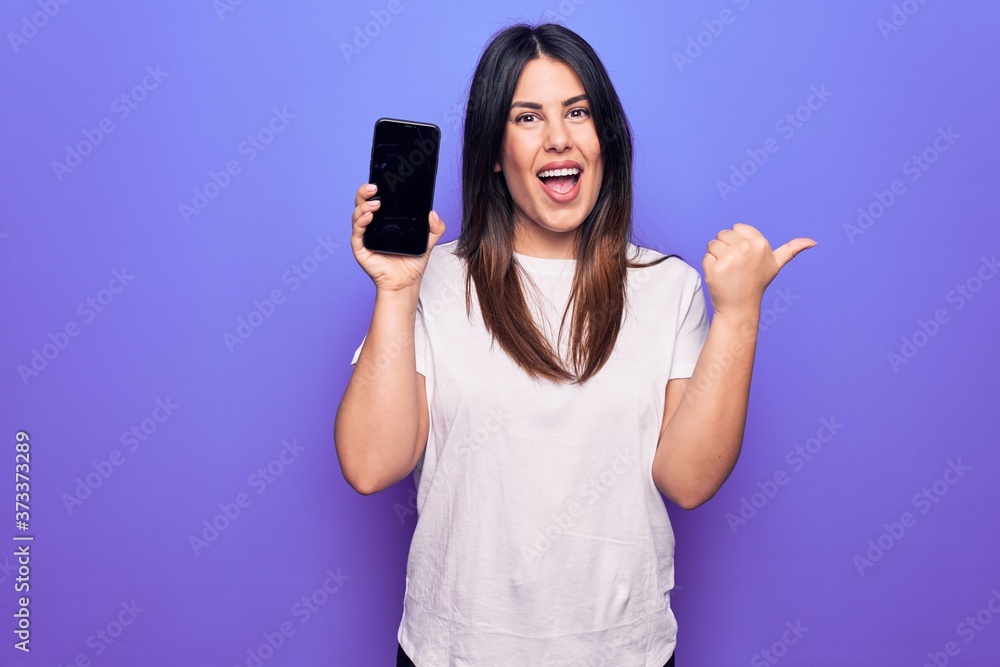 Young beautiful brunette woman holding smartphone showing screen over purple background pointing thumb up to the side smiling happy with open mouth Young beautiful brunette woman holding smartphone showing screen over purple background pointing thumb up to the side smiling happy with open mouth