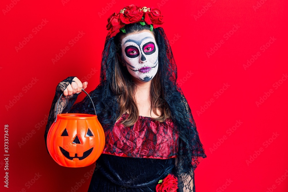 Young woman wearing day of the dead costume holding pumpkin thinking attitude and sober expression looking self confident Young woman wearing day of the dead costume holding pumpkin thinking attitude and sober expression looking self confident