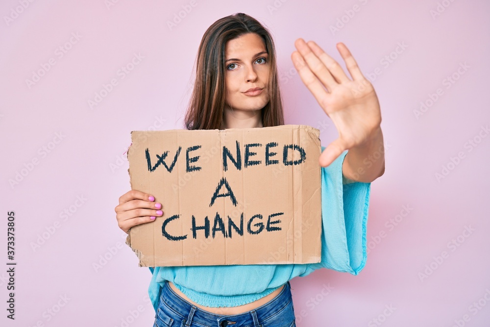 Beautiful caucasian woman holding we need a change banner with open hand doing stop sign with serious and confident expression, defense gesture Beautiful caucasian woman holding we need a change banner with open hand doing stop sign with serious and confident expression, defense gesture