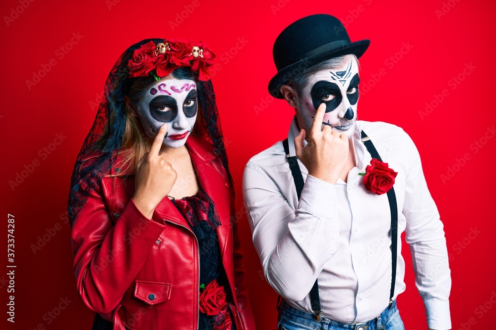 Couple wearing day of the dead costume over red pointing to the eye watching you gesture, suspicious expression Couple wearing day of the dead costume over red pointing to the eye watching you gesture, suspicious expression