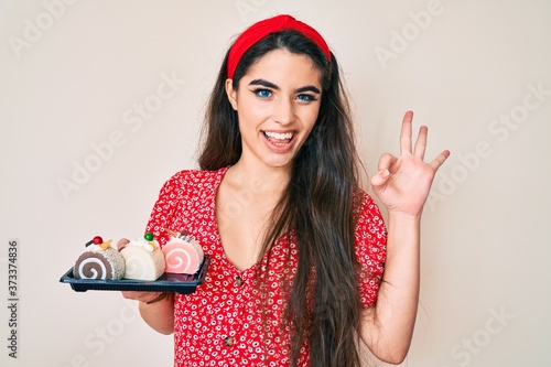 Brunette teenager girl holding cake sweets doing ok sign with fingers, smiling friendly gesturing excellent symbol