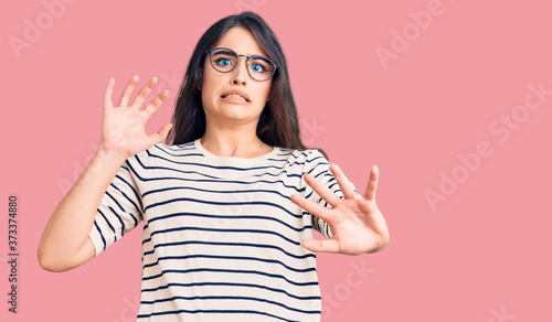 Brunette teenager girl wearing casual clothes and glasses afraid and terrified with fear expression stop gesture with hands, shouting in shock. panic concept. © Krakenimages.com