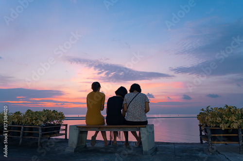Three asian girls sit on a bench by the sea and watch the sunset
