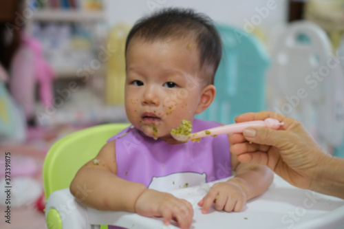 Mother feeding the baby's meal with a spoon. 9 months old Asian baby eat food in the chair for baby. Selected focus. Vintage tone.