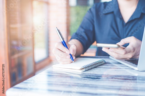 Woman is writing on a notebook with a pen and she is using a mobile phone in the office.