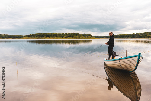 An elderly man is fishing from a boat near the shore on the lake © Sergey + Marina