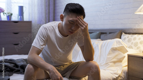 portrait of man sitting at bedside touching forehead is suffering from insomnia. asian male staying awake until daybreak is painful. sleep disorder and health problem concept. photo