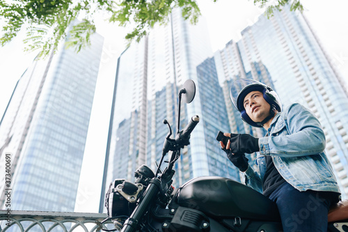 Handsome serious Vietnamese young man in helmet sitting on motorcycle with smartphone in hands, skyscrapers in background