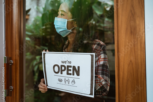 asian woman with open sign in her shop during covid-19 pandemic. new normal life keep your distance sign
