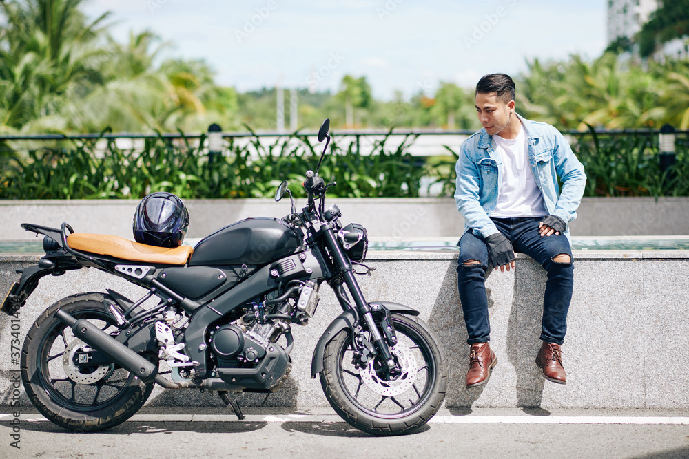 Proud Vietnamese biker sitting on parapet and looking at his motorcycle