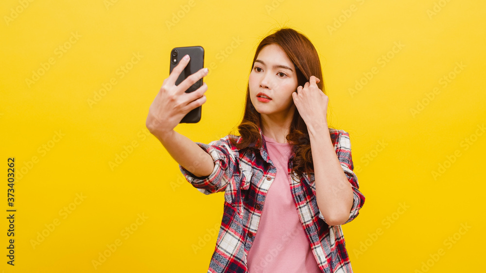 Smiling adorable Asian female making selfie photo on smartphone with positive expression in casual clothing and looking at camera over yellow background. Happy adorable glad woman rejoices success.