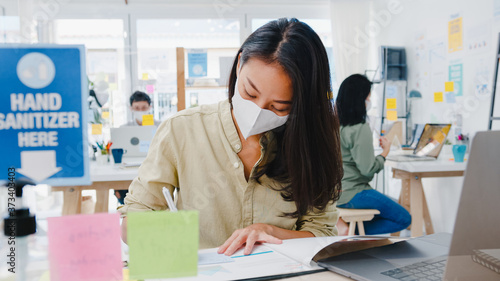 Asia businesswoman entrepreneur wearing medical face mask for social distancing in new normal situation for virus prevention while using laptop back at work in office. Life and work after coronavirus.