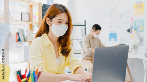 Asia female wear face mask for social distancing in new normal situation for virus prevention while using laptop and separated by acrylic partition stand in office. Life and work after corona virus.