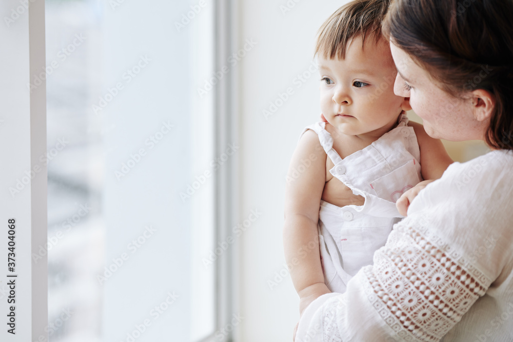 Mother carrying her little daughter when they are standing at window and looking outside Mother carrying her little daughter when they are standing at window and looking outside