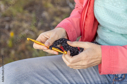 Needlework. A girl in jeans and a red jacket is crocheting a black rug from a thick cord in nature.