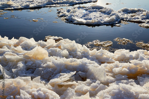 Ice drift on a river with blue high water and big water, white snow broken ice full of hummocks in it and sun reflection in sunny spring day.