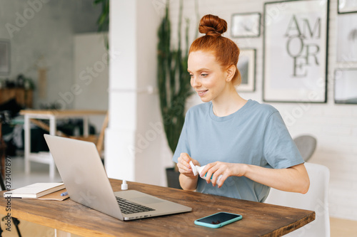 Cute young redhead businesswoman is wiping laptop computer with sanitizer before starting work in light cozy room at the home office, looking at display screen. Concept of distance job on Internet. 