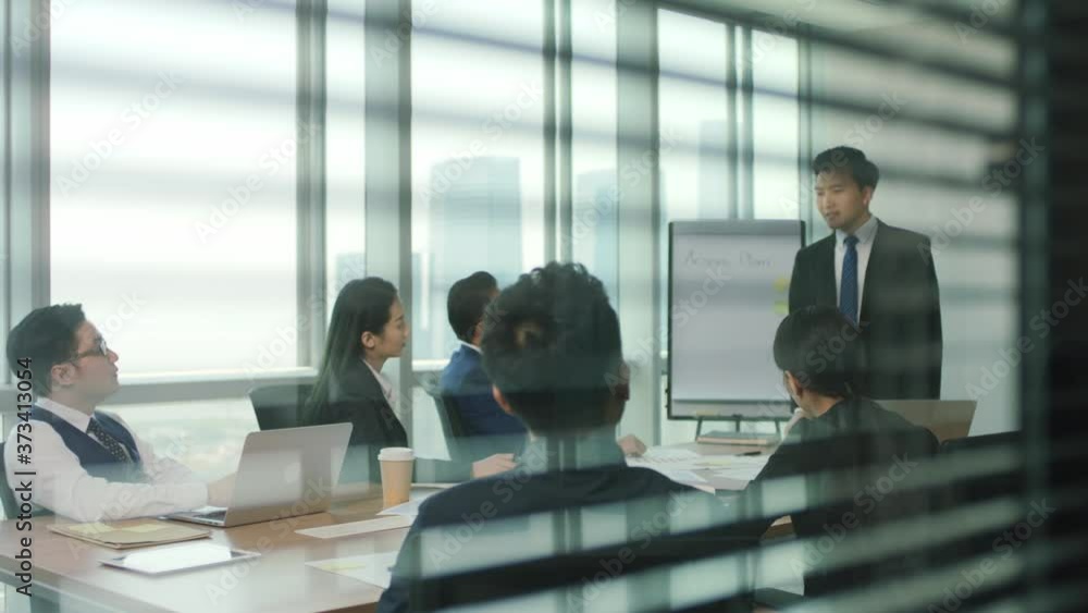 through the glass and blinds shot of a team of asian business people meeting in modern office conference room