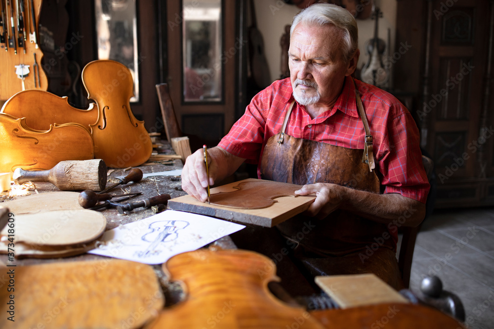 Arts and crafts. Senior carpenter craftsman making violin music instrument in his old-fashion carpenter's workshop. In background hardwood violin parts and tools. Arts and crafts. Senior carpenter craftsman making violin music instrument in his old-fashion carpenter's workshop. In background hardwood violin parts and tools.