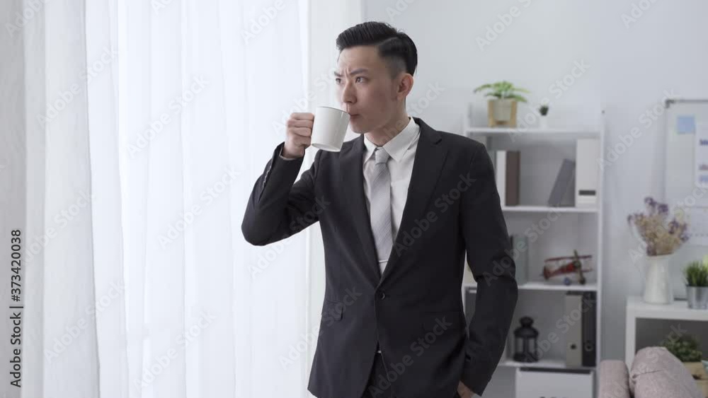 male ceo is sipping morning coffee by window with serious expression. asian man in black suit with hand in pocket is looking outside in contemplation male ceo is sipping morning coffee by window with serious expression. asian man in black suit with hand in pocket is looking outside in contemplation