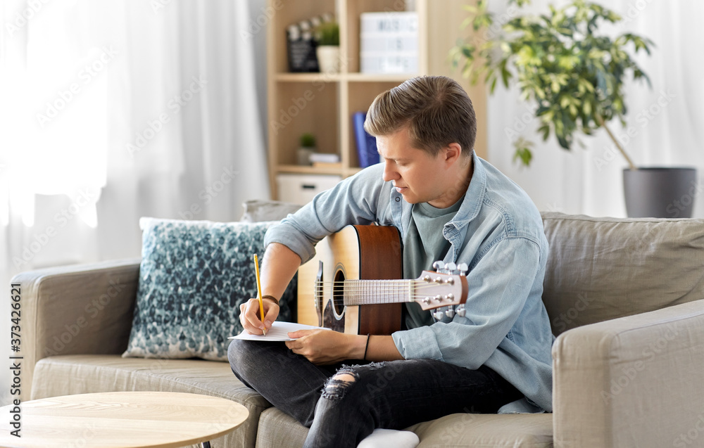 leisure and people concept - young man or musician with guitar sitting on sofa and writing to music book at home