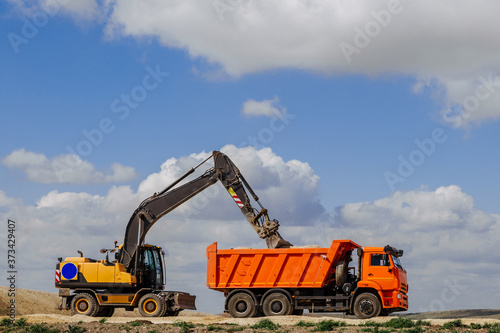 A yellow backhoe loader loads the earth into a truck during the construction of a road against a blue sky with clouds.