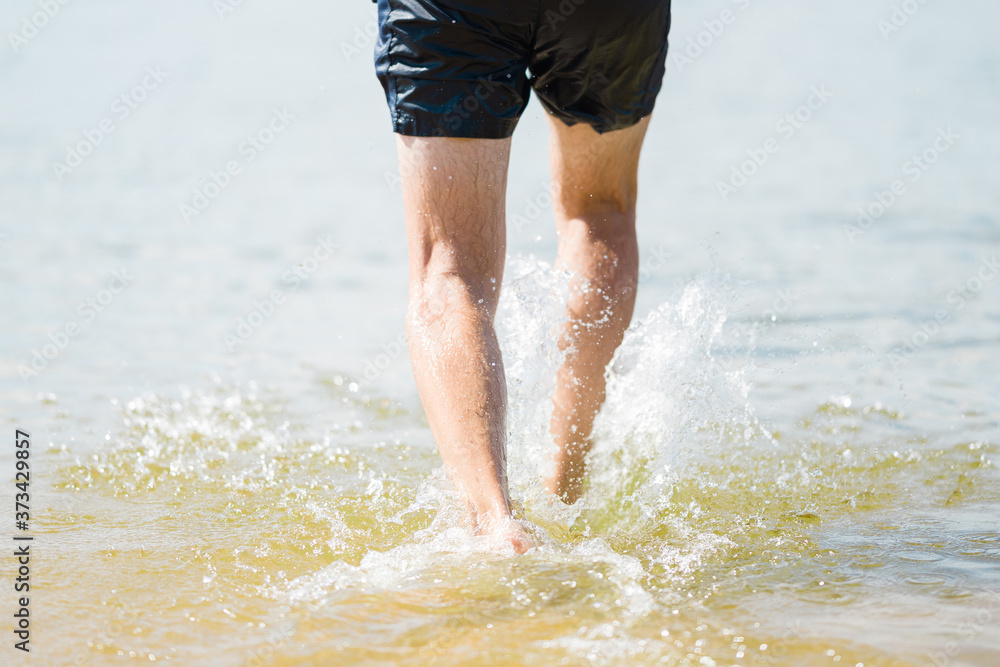 Young man legs walking in sea water in sunny day. Back view. Closeup.