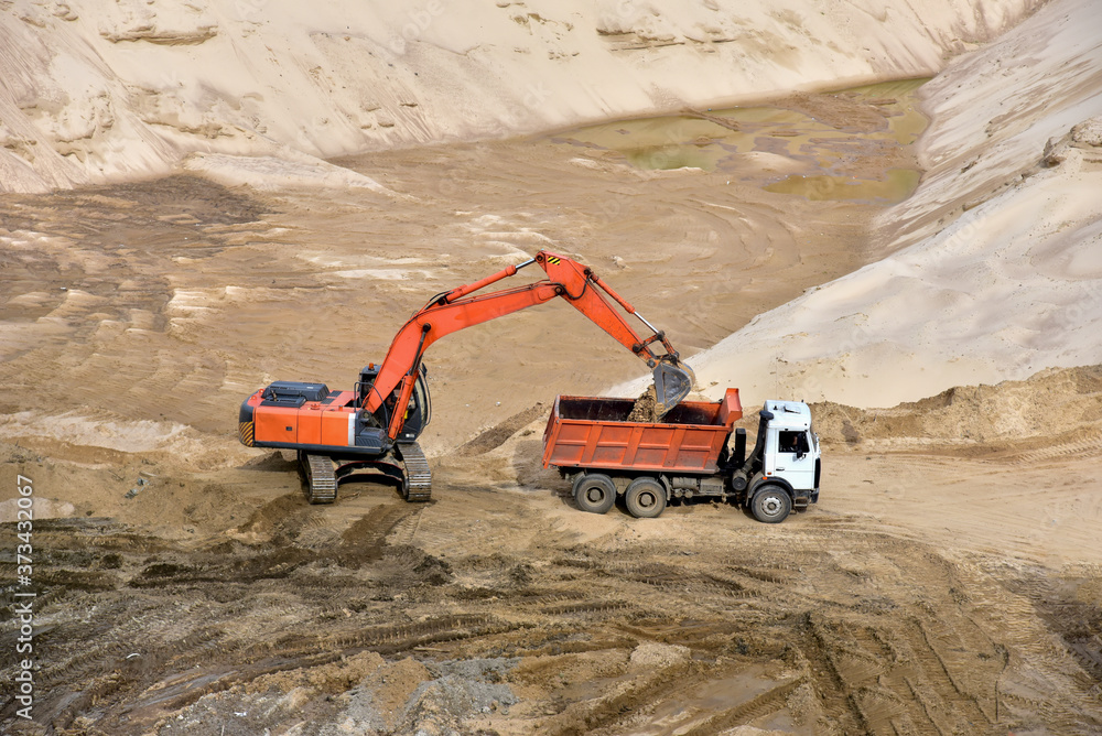 Excavator load the sand to the heavy dump truck in the open-pit. Heavy machinery working in the mining quarry. Digging and excavation operations. View on the mountains with yellow sand. Excavator load the sand to the heavy dump truck in the open-pit. Heavy machinery working in the mining quarry. Digging and excavation operations. View on the mountains with yellow sand.