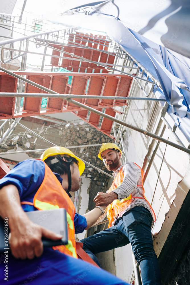 Builder in vest and hardhat helping coworker to climb up the scaffolding outside the building under construction Builder in vest and hardhat helping coworker to climb up the scaffolding outside the building under construction