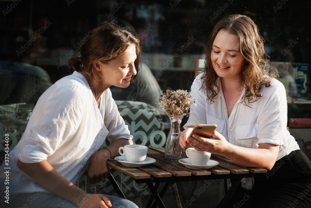 Two young females using smartphone in a cafe outdoors sitting in a cafe with coffee Two young females using smartphone in a cafe outdoors sitting in a cafe with coffee